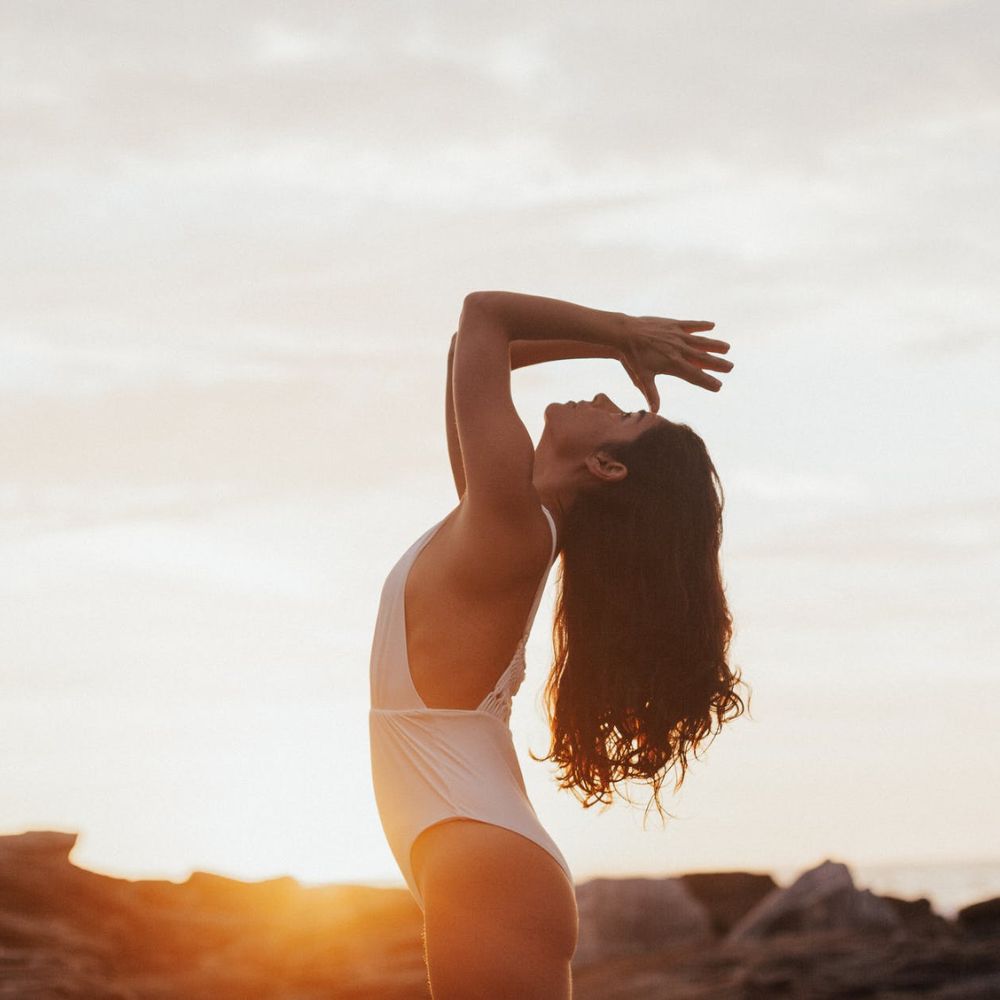 Bài tập trị đau cổ - Cô gái trong trang phục tập yoga màu trắng đang thực hiện động tác kéo giãn cổ dưới ánh hoàng hôn