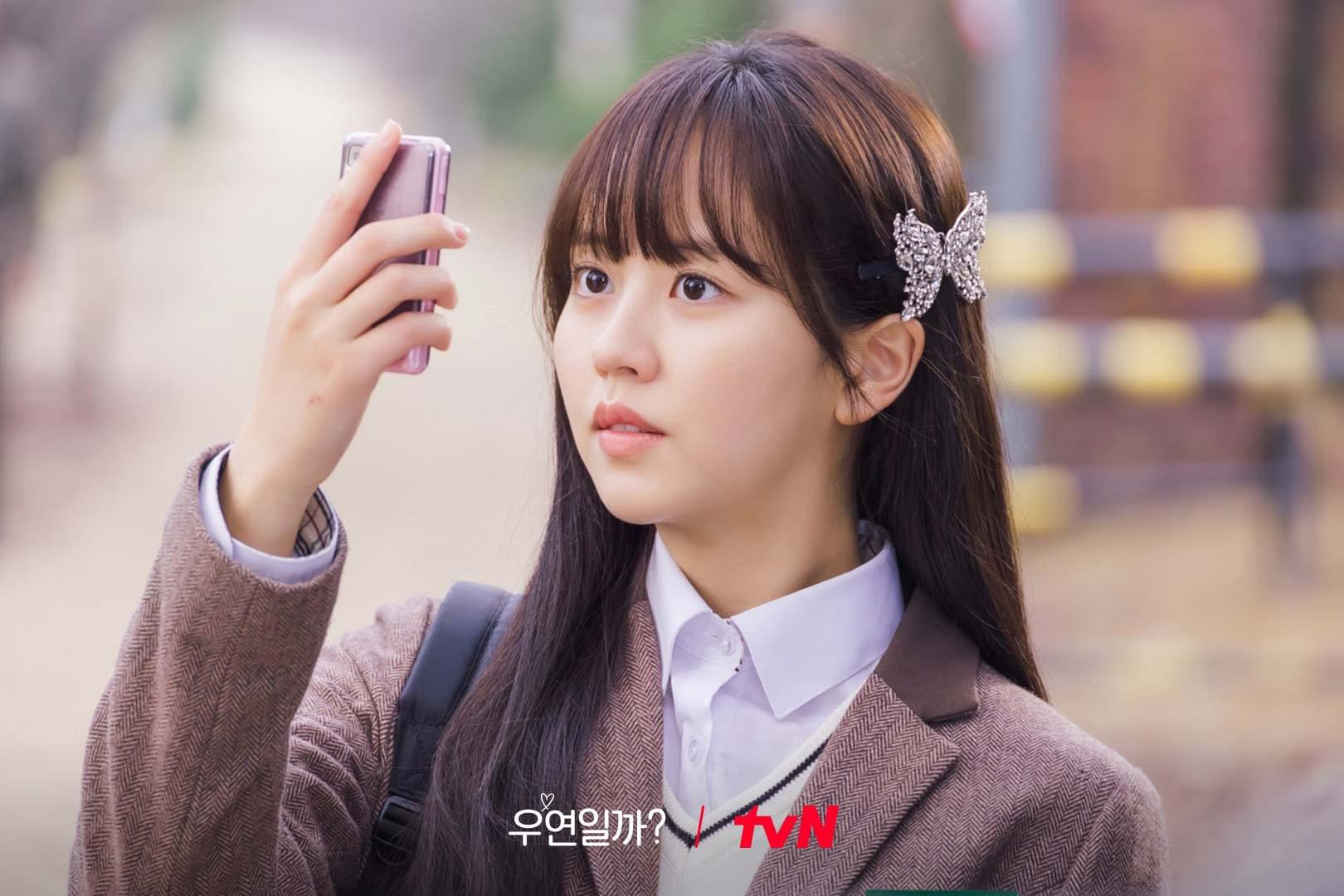 Kim So Hyun mặc đồng phục học sinh màu nâu, kẹp tóc hình bướm và cầm điện thoại selfie