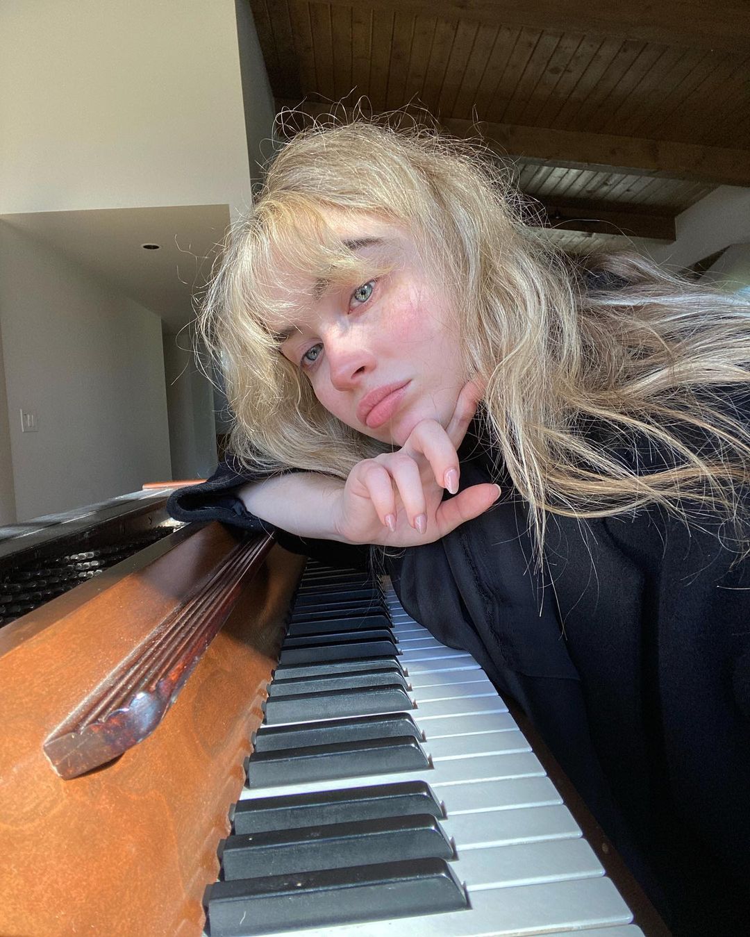 Sabrina Carpenter mặc áo thun màu đen, nhuộm tóc bạch kim và tạo dáng nằm nghiêng bên đàn piano