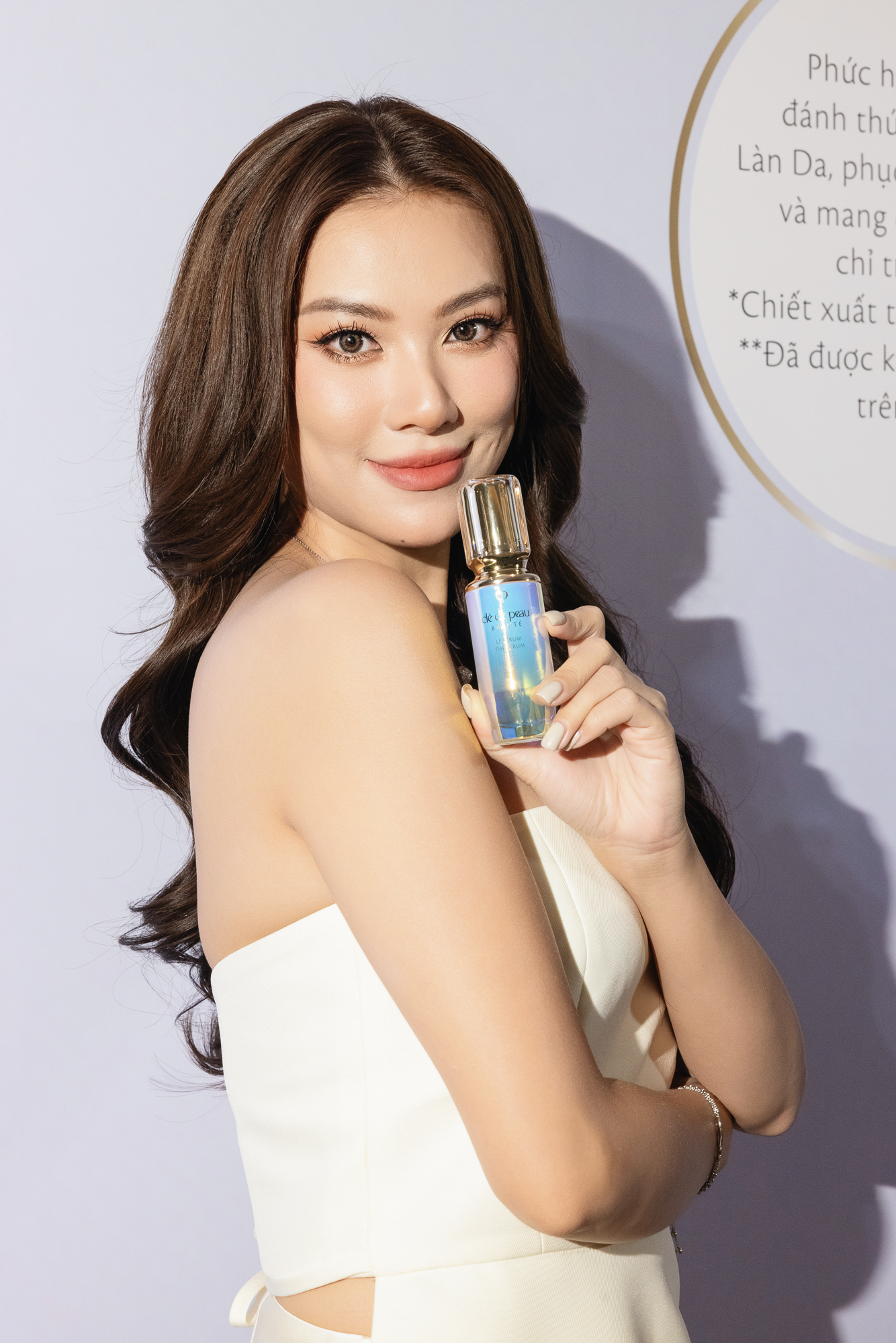 serum - á hậu Kim Duyên mặc đầm dự tiệc màu trắng, tạo kiểu tóc uốn xoăn và cầm chai tinh chất The Serum của Cle de Peau để chụp ảnh chân dung